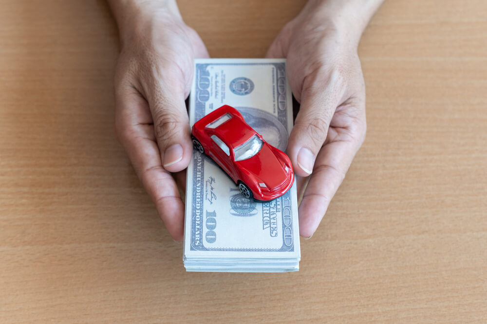  A hand holding a bundle of $100 cash with a red toy car on top of it on a brown wooden table.