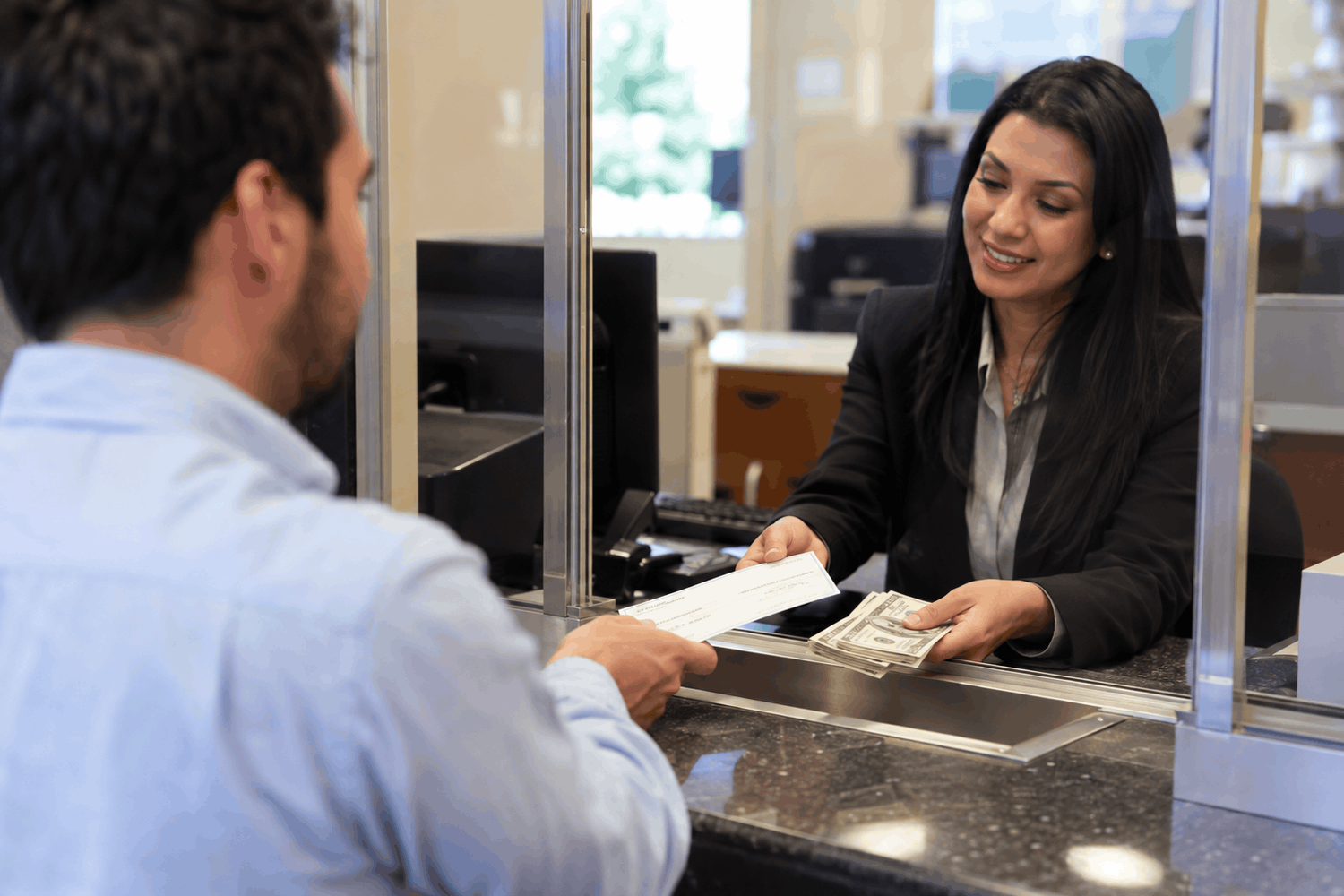 A customer cashing a check at a bank counter while a teller hands over cash through a glass service window.