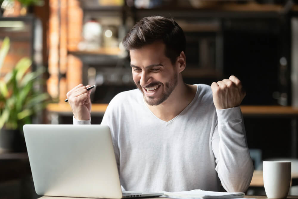 A man excited about online title pawn in a room with a laptop, flower vase, and a cup of coffee on the table.