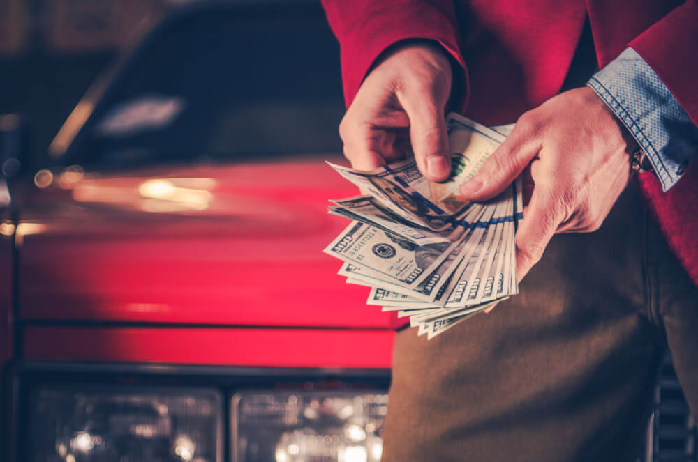 Fast cash title loan money is highlighted in the image. A man wearing red is shown holding a $100 bill in his hand. In the background, there is a red car.