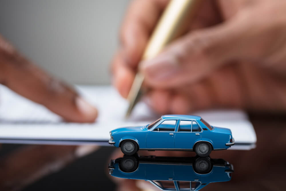 The image portrays someone signing an auto pawn agreement, accompanied by an image of a toy car, symbolizing a collateral asset.