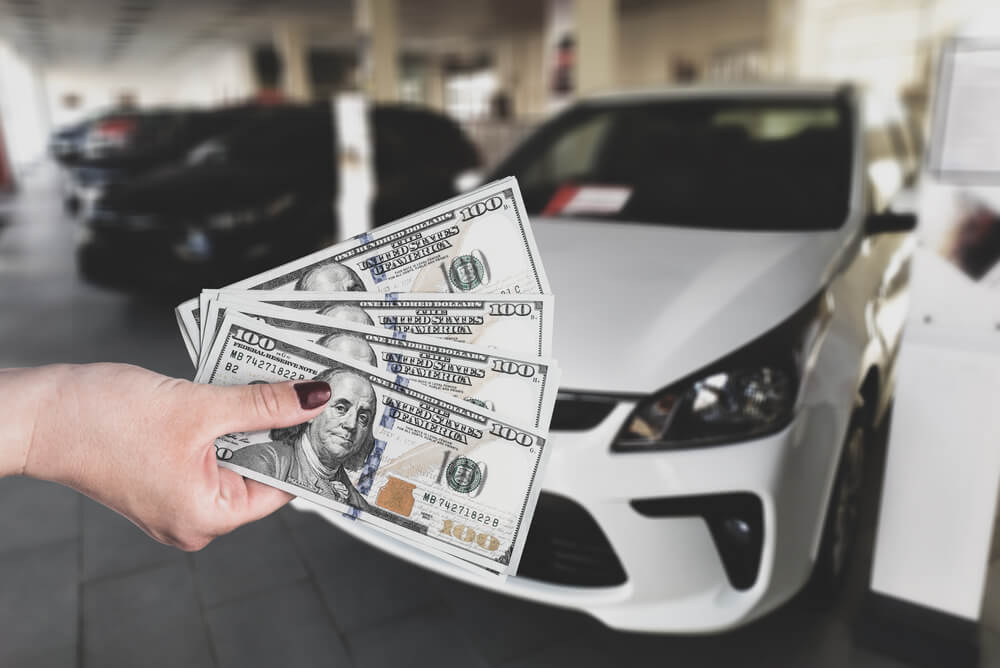 Depicts a car title loans A hand holding a 400$ cash in a parking lot with white and black card in a background.