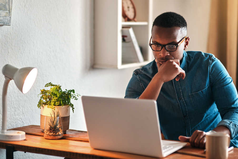  A man in a room, wearing eyeglasses, is sitting in a chair at a table with a laptop, a glass, and a flower vase.