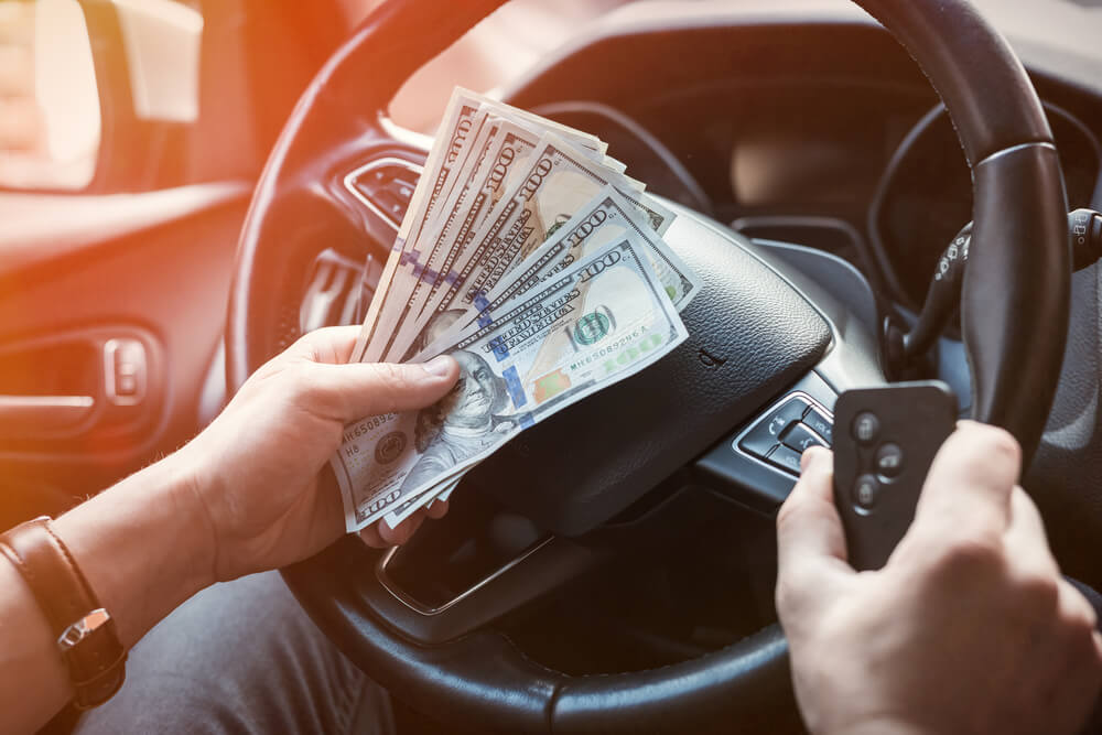 Image shows a man inside car holds auto title loan cash, steering wheel visible.